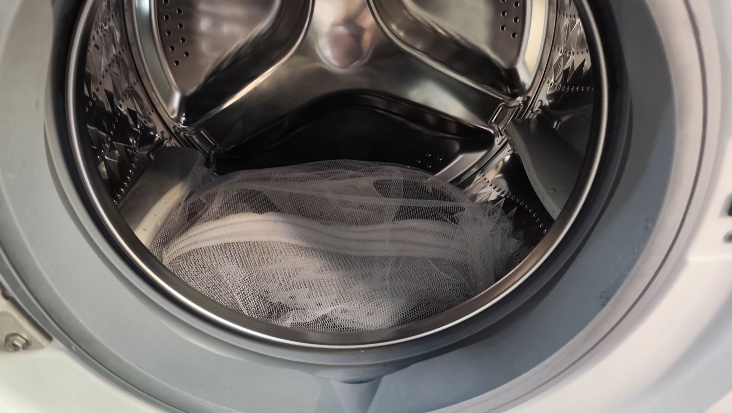 Inside view of washing machine drum showing grey sneakers in white mesh laundry bag ready to wash