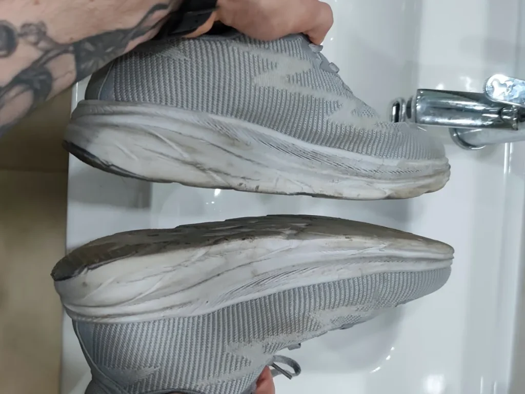Hands holding grey sneakers over bathroom sink knocking soles together to remove loose dirt