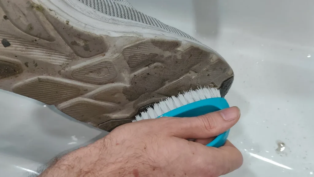 Hand using blue scrub brush to clean dirty rubber outsole of grey running shoe
