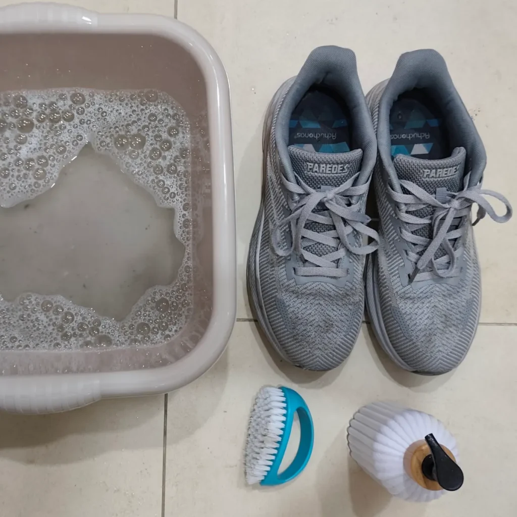 Flat lay of grey sneakers with washing basin soapy water blue brush and toothbrush on tiled floor