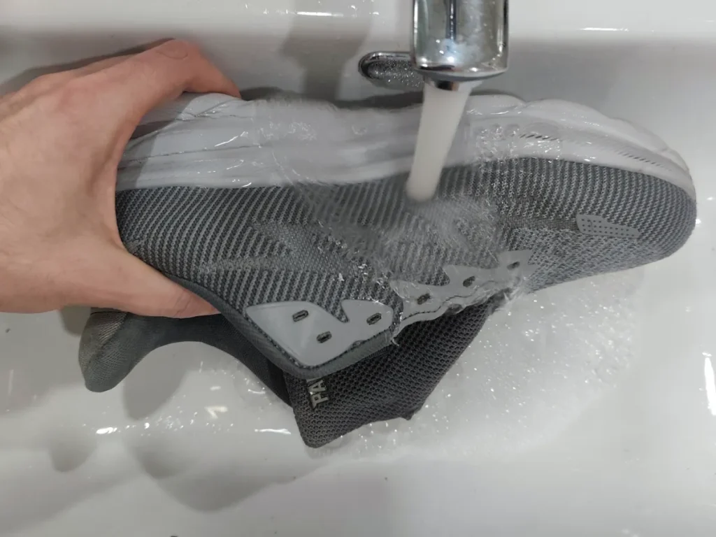 Hand holding grey mesh sneaker under running cold water tap to rinse off soap after hand washing