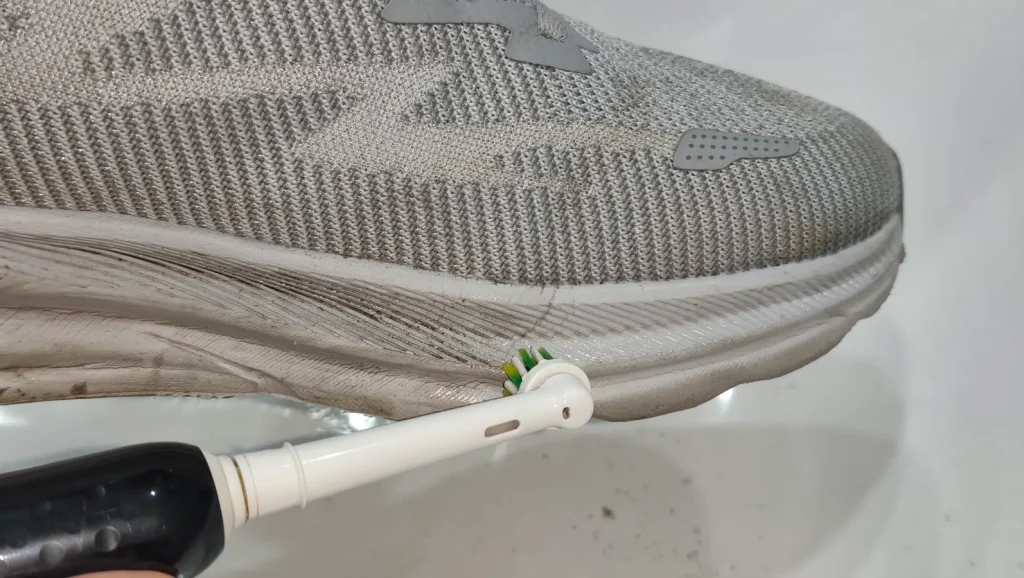 Hand holding toothbrush scrubbing dirt from grey sneaker midsole edge in bathroom sink